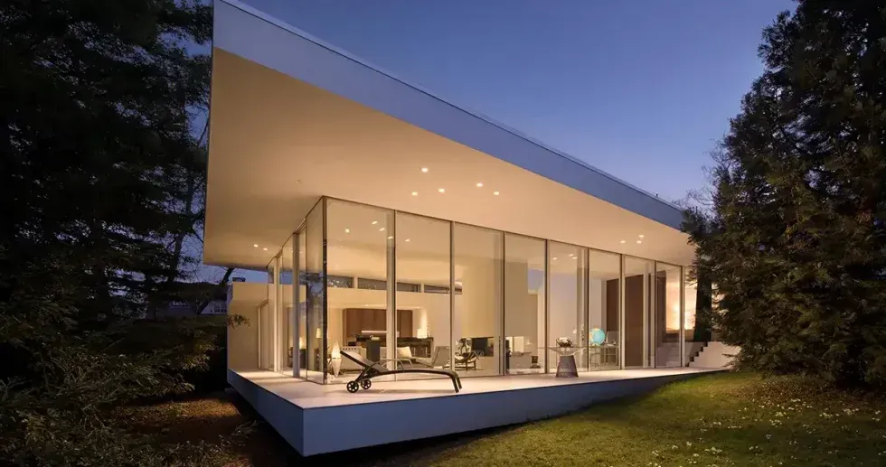 Modern glass house with a slanted roof, lit interior, and a forested outdoor setting.