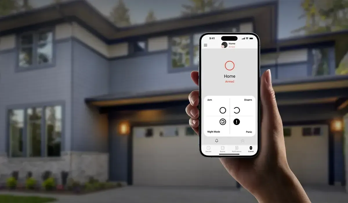 Hand holding a smartphone showing a home security app interface in front of a modern two-story house.