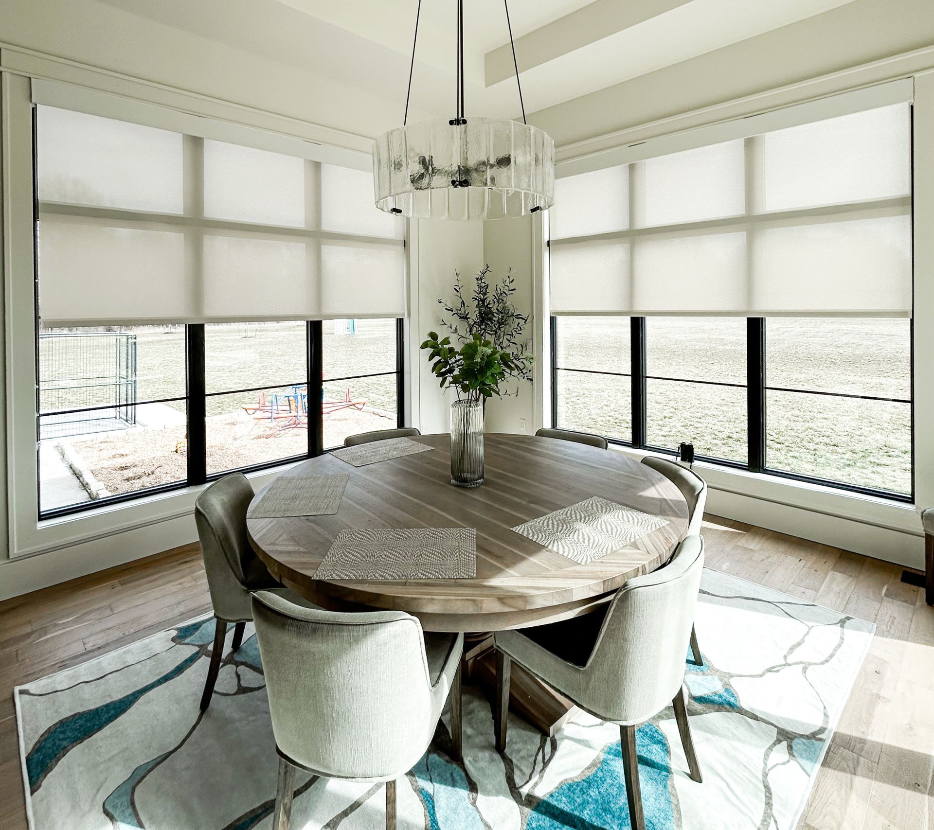 Dining room with a round wooden table, chairs, and large windows with blinds.