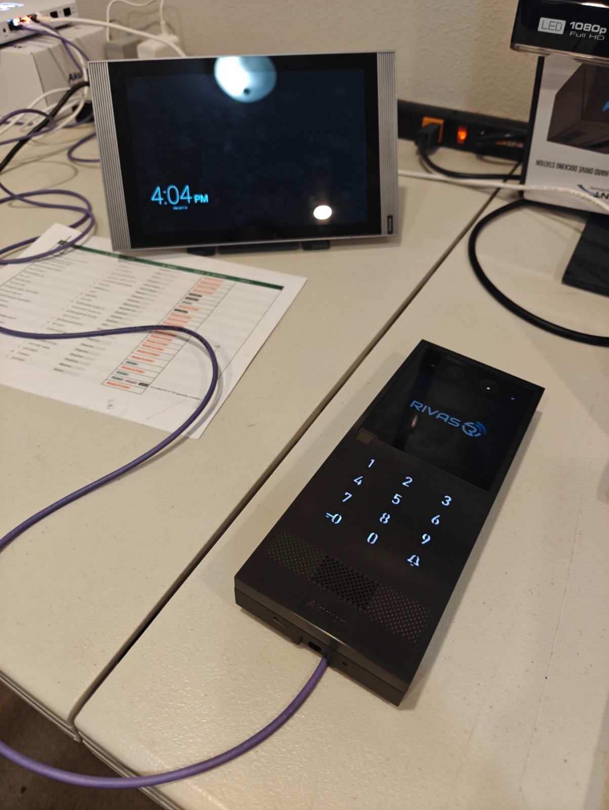 A black keypad device with a small screen sits next to a tablet on a table; both have displays. A cable connects the keypad.