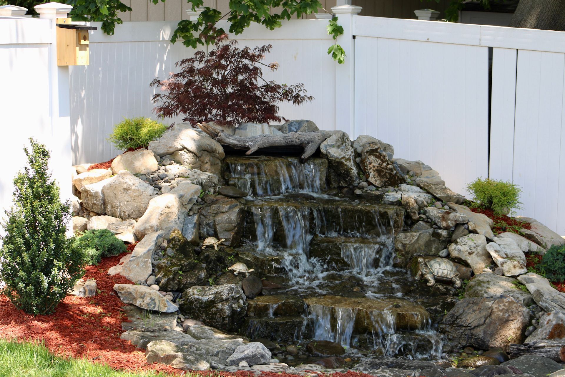 A waterfall in a backyard with a white fence in the background