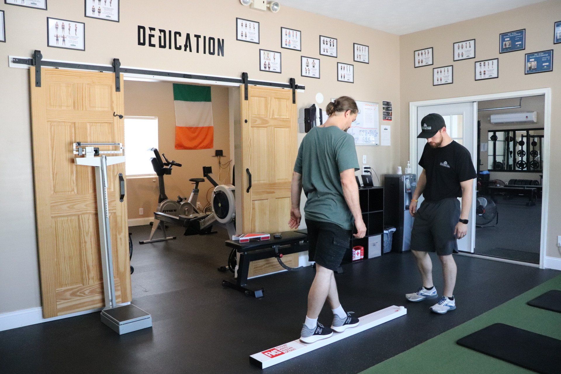 Two people standing in a gym near a bench and weightlifting equipment, with “DEDICATION” on the wall.