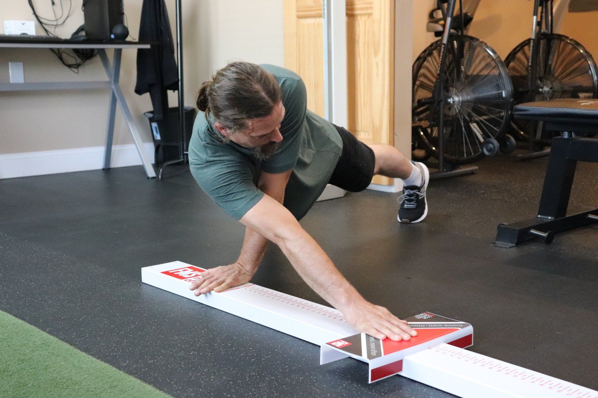 Man balancing on a white board with arms outstretched in a gym, reaching toward the floor