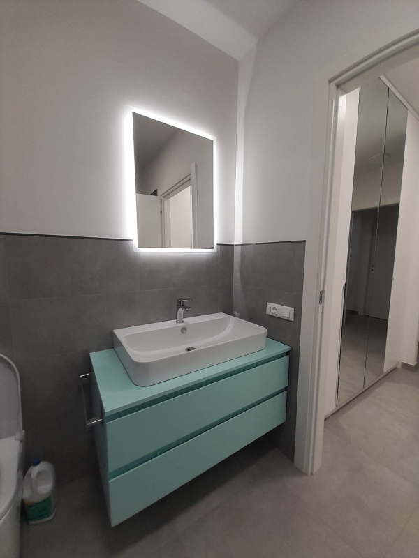 Un bagno moderno con mobiletto azzurro, lavabo a ciotola bianco e specchio quadrato illuminato su pareti rivestite di piastrelle grigie.
