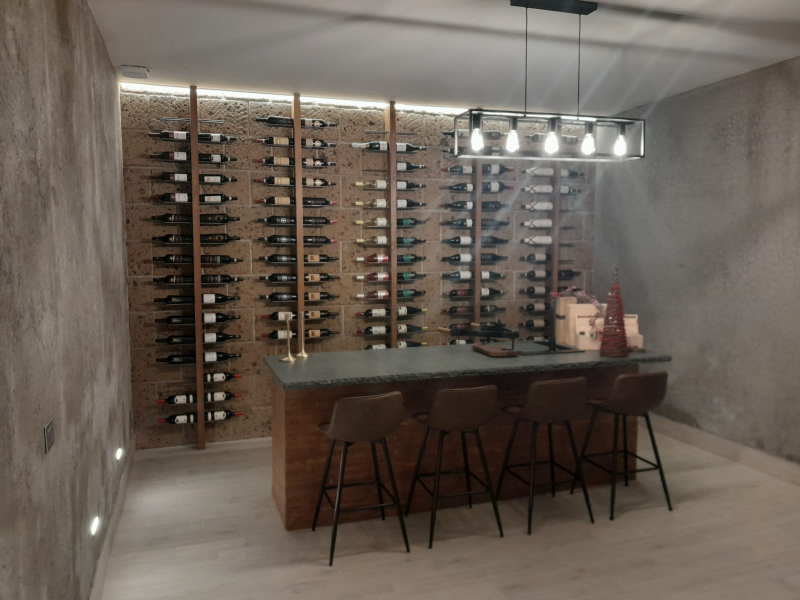 Una cantina domestica con scaffalature per bottiglie a tutta altezza, un'isola bar in legno, quattro sgabelli e lampade a sospensione in stile industriale.