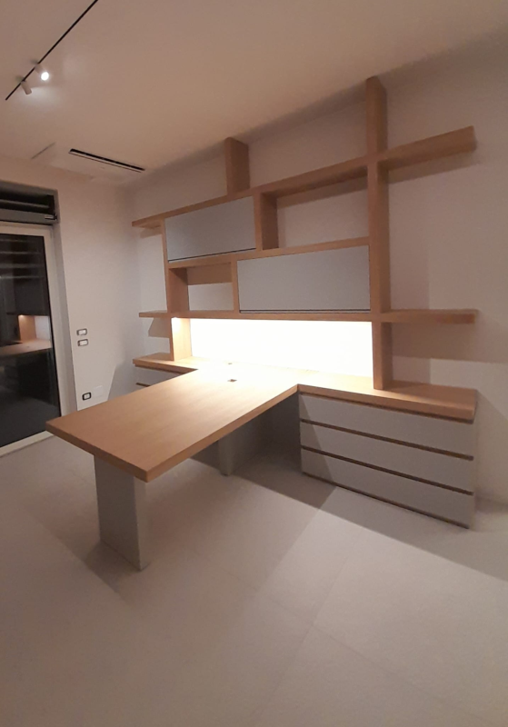 Un moderno ufficio domestico con scrivania in legno chiaro, cassetti e mensole asimmetriche a parete illuminate da luci integrate.