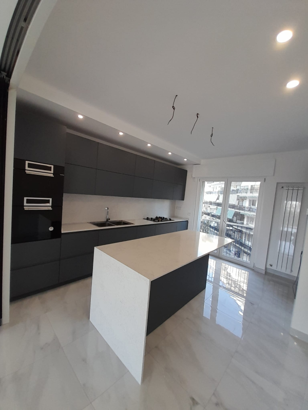 Una cucina moderna con mobili grigio scuro opaco, un'isola in quarzo bianco e pavimento lucido di colore chiaro.