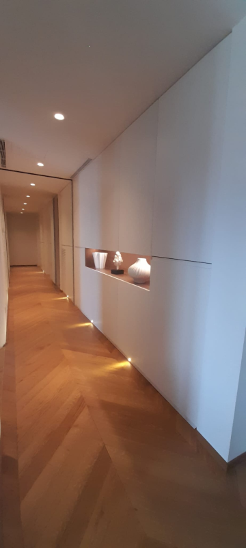 Un corridoio luminoso con pavimento in legno chiaro a spina di pesce, pannelli a parete bianchi e una mensola incassata e illuminata con oggetti decorativi.