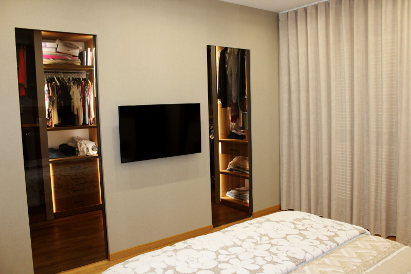 Una camera da letto con un televisore montato a parete tra due armadi alti e illuminati con ante in vetro, accanto a tende beige.