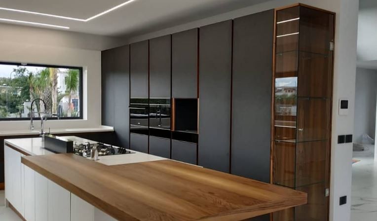Cucina moderna con isola bianca, bancone per la colazione in legno e mobili a tutta altezza di colore grigio scuro con vetrina in legno.