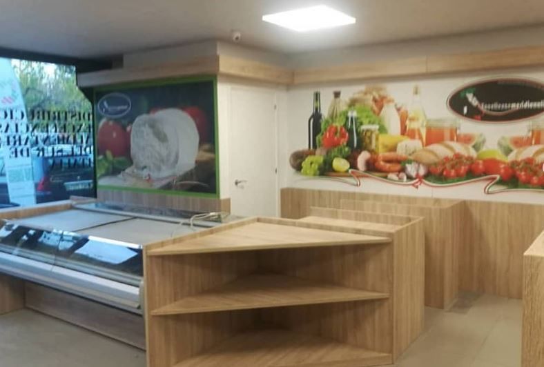 Un negozio d'angolo con banconi in legno chiaro, una vetrina refrigerata e murales alle pareti raffiguranti prodotti alimentari.