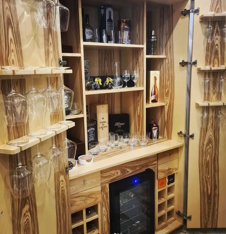 Un mobile bar in legno per la casa, con bicchieri da vino appesi, ripiani per bottiglie di liquore e un frigorifero per il vino integrato.
