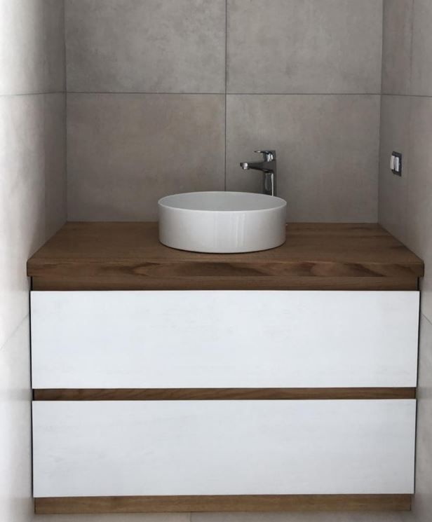 In un bagno, un lavabo a ciotola bianco è appoggiato su un mobiletto in legno con due cassetti bianchi, contro una parete rivestita di piastrelle grigie.