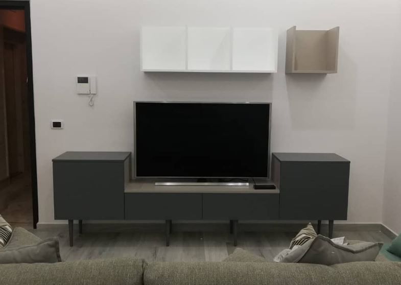 Un mobile porta TV grigio scuro con televisore, sopra il quale si trovano pensili bianchi e una mensola quadrata aperta su una parete bianca.