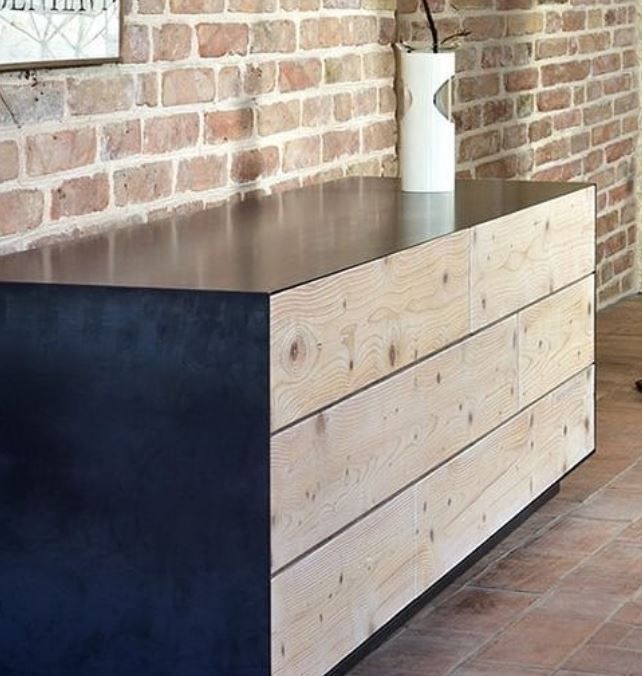 Una cassettiera moderna con fianchi in metallo scuro e frontali dei cassetti in legno chiaro nodoso, incastonata contro una parete di mattoni rustici.