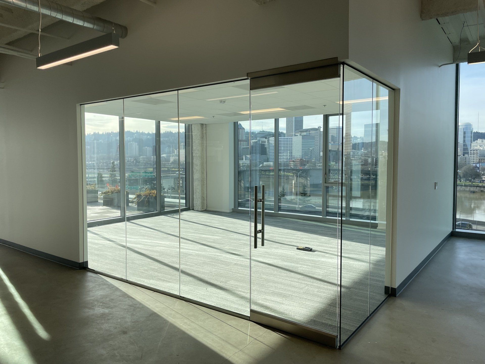 An empty office conference room enclosed by glass walls, featuring a glass door, carpeted floor, and city view outside.