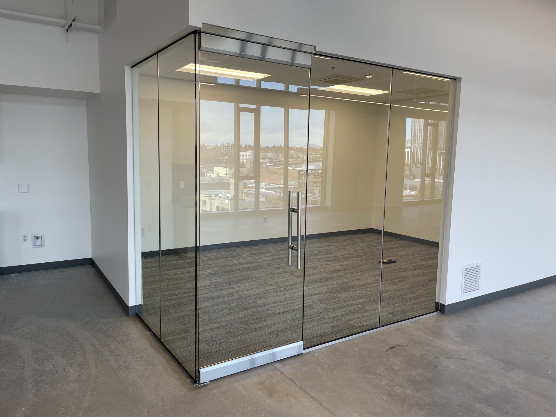 A modern, glass-walled office conference room with a door, metal frames, and grey carpet, set in an open workspace.