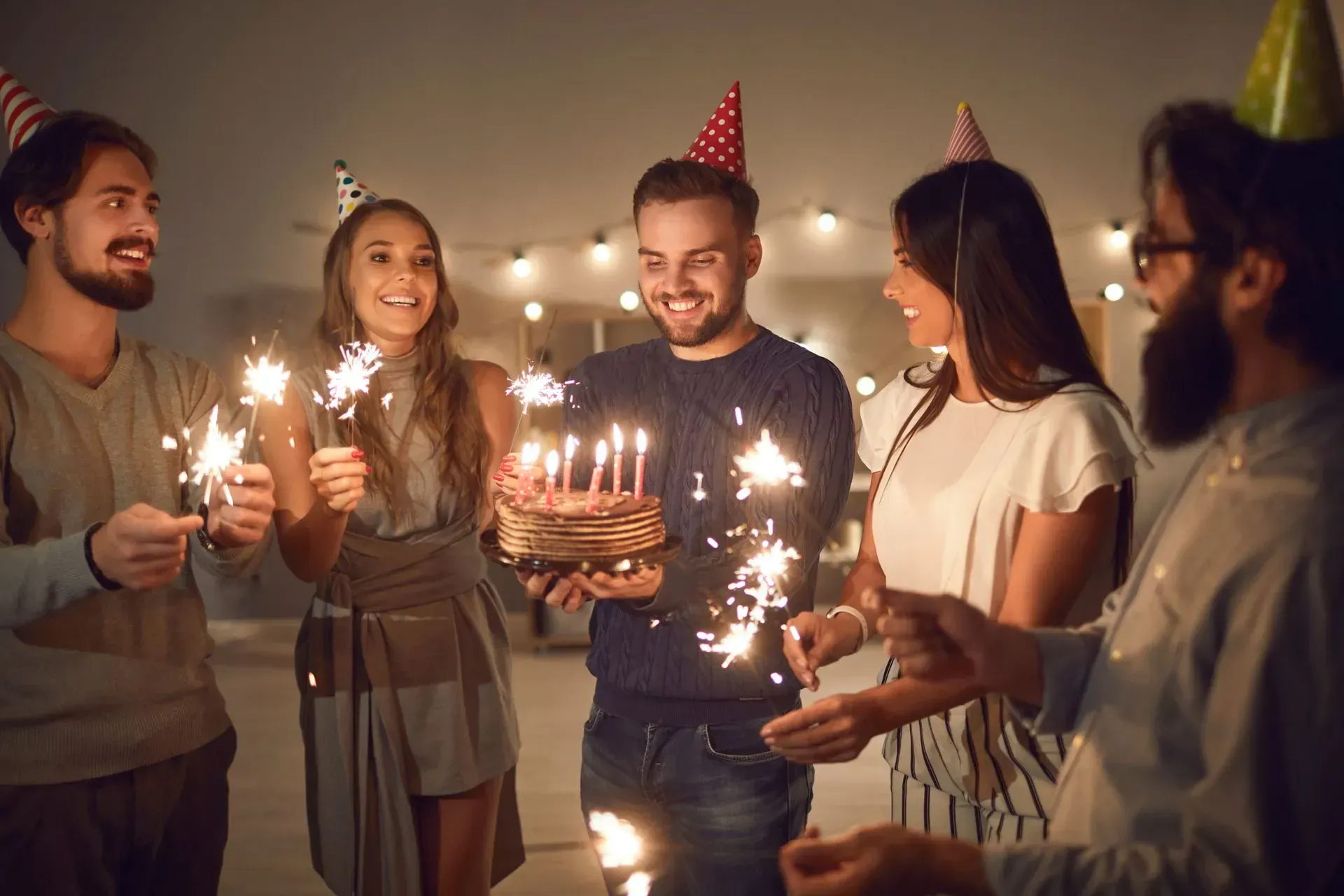 Friends in party hats hold sparklers while a person holds a birthday cake with lit candles in a softly lit room.