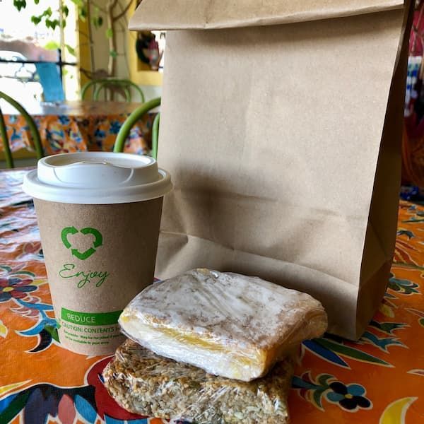 A paper cup that says enjoy next to a to go paper bag with an organic lemon bar and fig bar in front of it.
