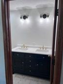 A bathroom vanity with a white countertop, two gold faucets, black cabinets, and two wall sconces viewed through a doorway.