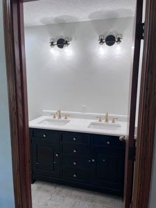 A bathroom vanity with a white countertop, two gold faucets, black cabinets, and two wall sconces viewed through a doorway.