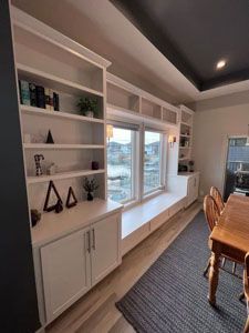 White custom built-in bookshelves and storage cabinets framing a large window in a dining room with a wooden table.