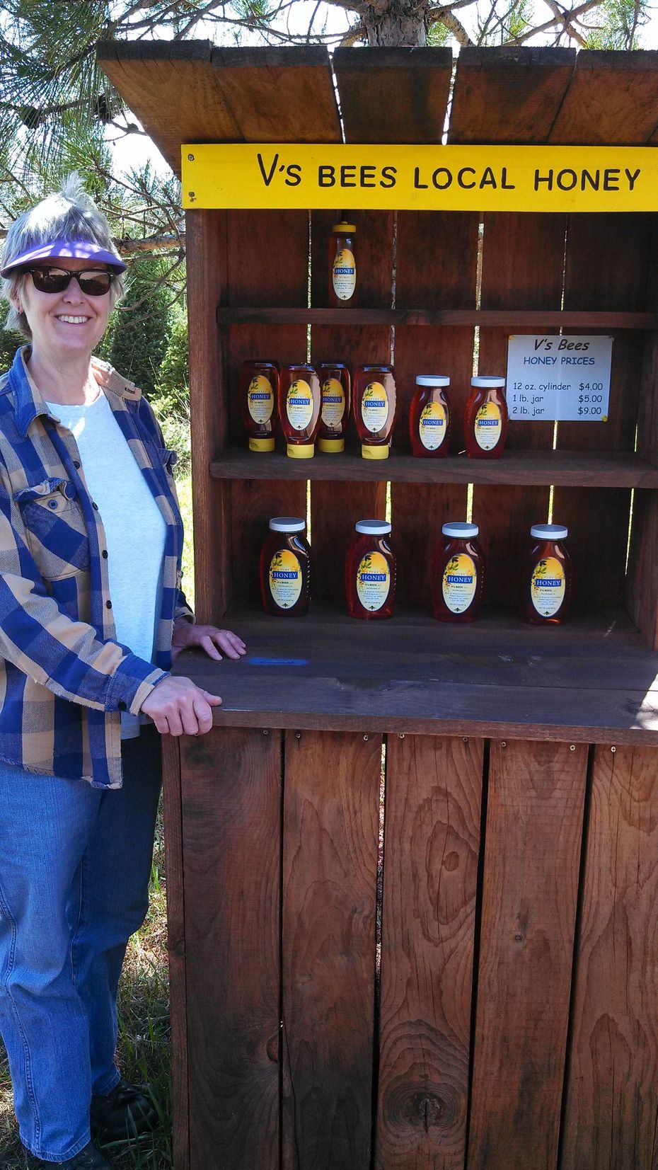 Woman beside honey stand with jars for sale under a yellow “V3 Bees Local Honey” sign