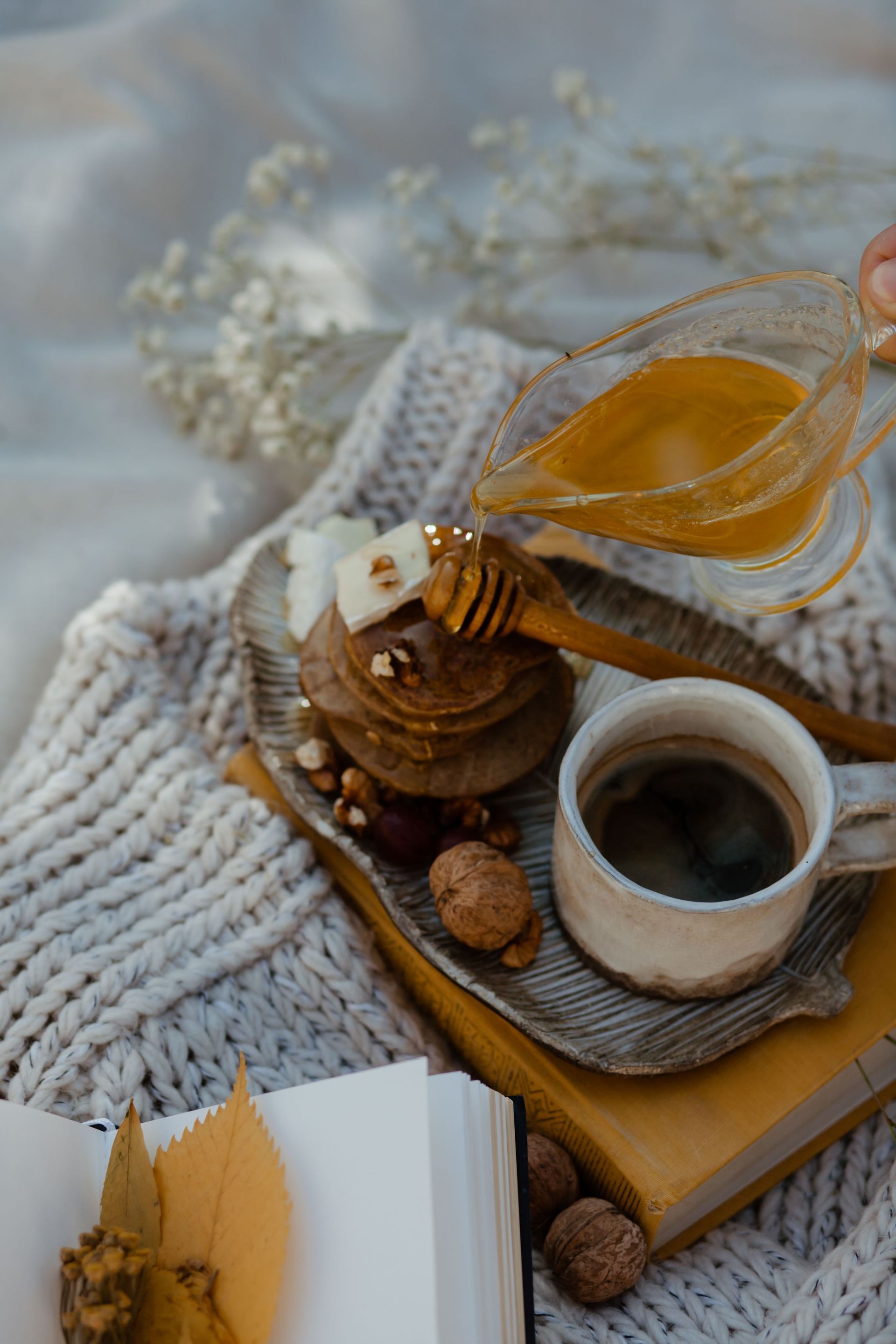 Cozy tea-and-breakfast tray with honey, coffee, pastries, and a knitted blanket in soft neutral tones