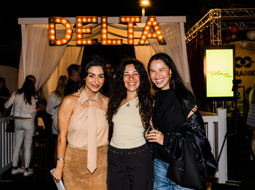 Three women smiling at a DELTA event, standing together indoors with warm marquee lights behind them.