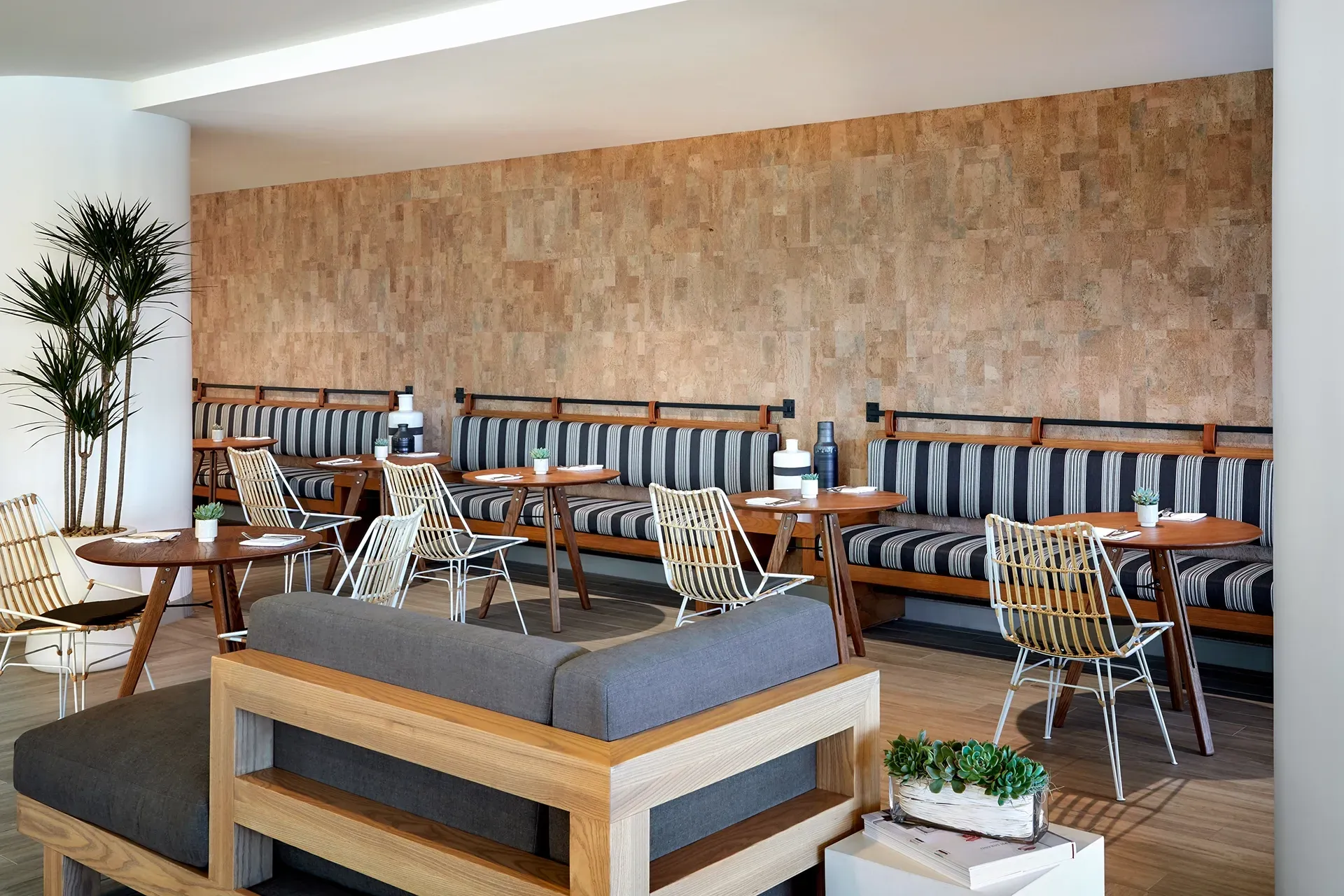 Modern lounge with wooden tables, woven chairs, and a gray sofa against a textured wall.