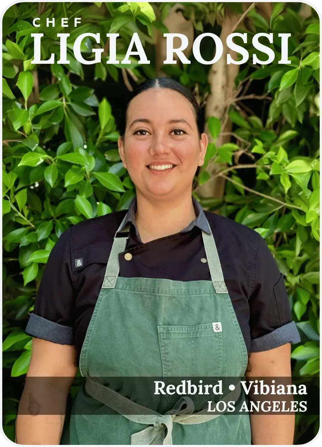 Chef Ligia Rossi smiling in a black chef coat and green apron, standing before a leafy green wall.