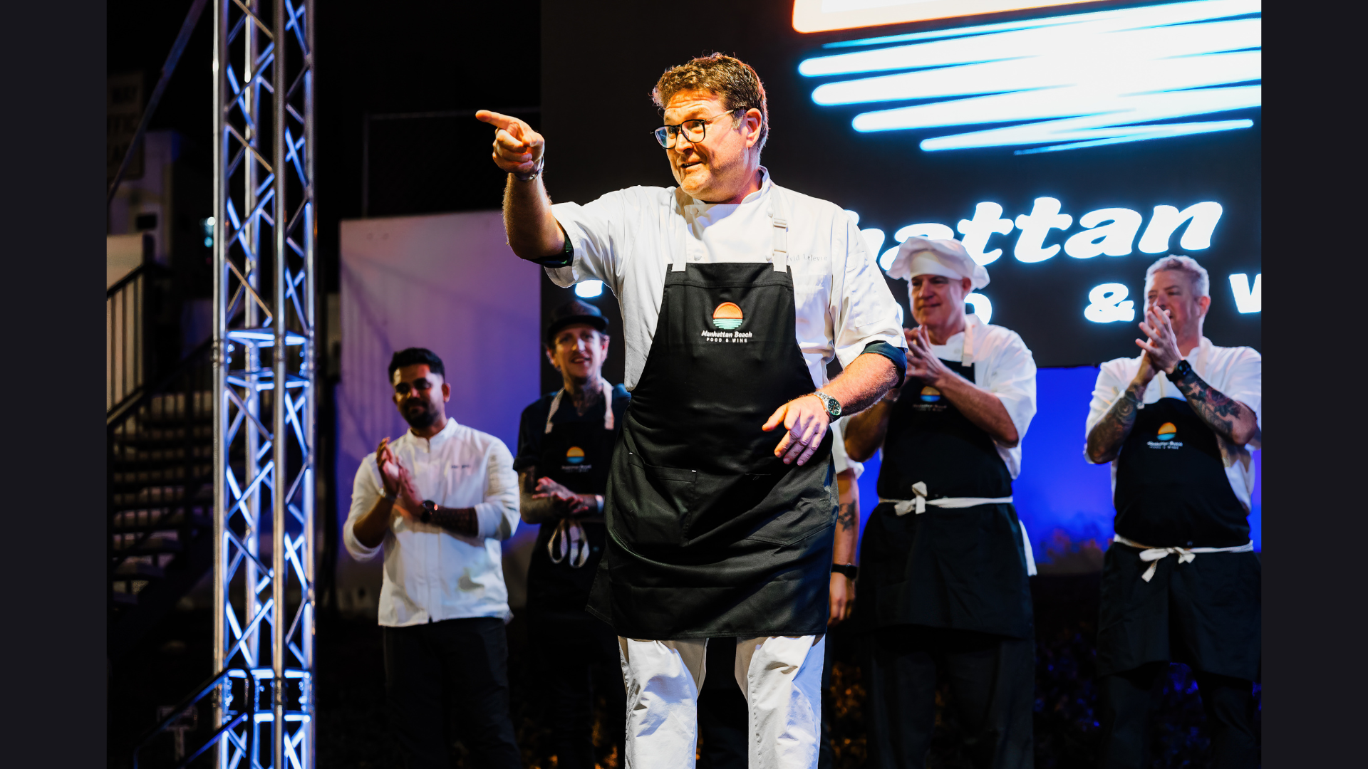 Chef in black apron gesturing on stage while others clap behind him under bright lights