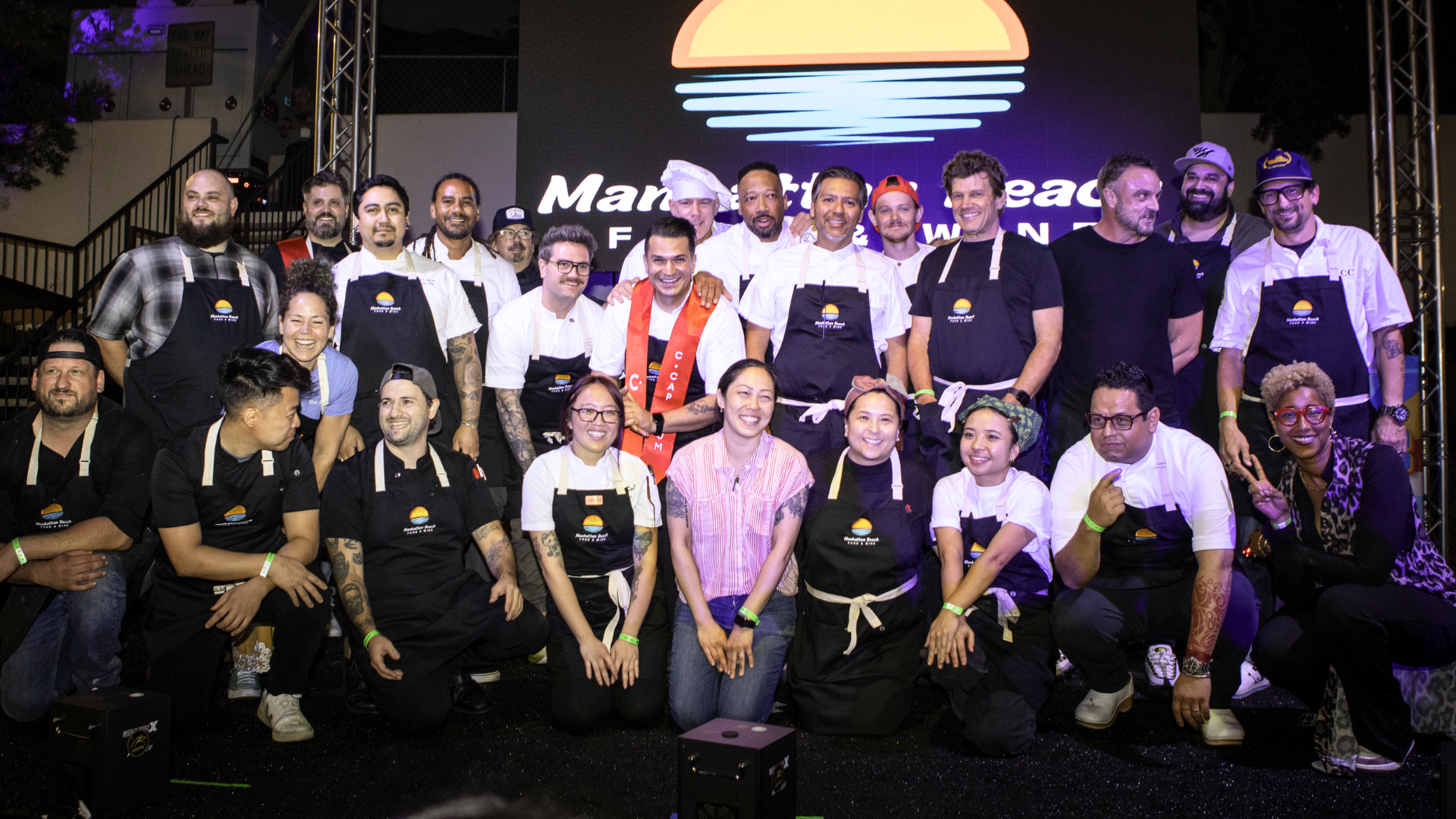 Group of people posing on a stage in front of a glowing sunset logo, many wearing black shirts and medals.