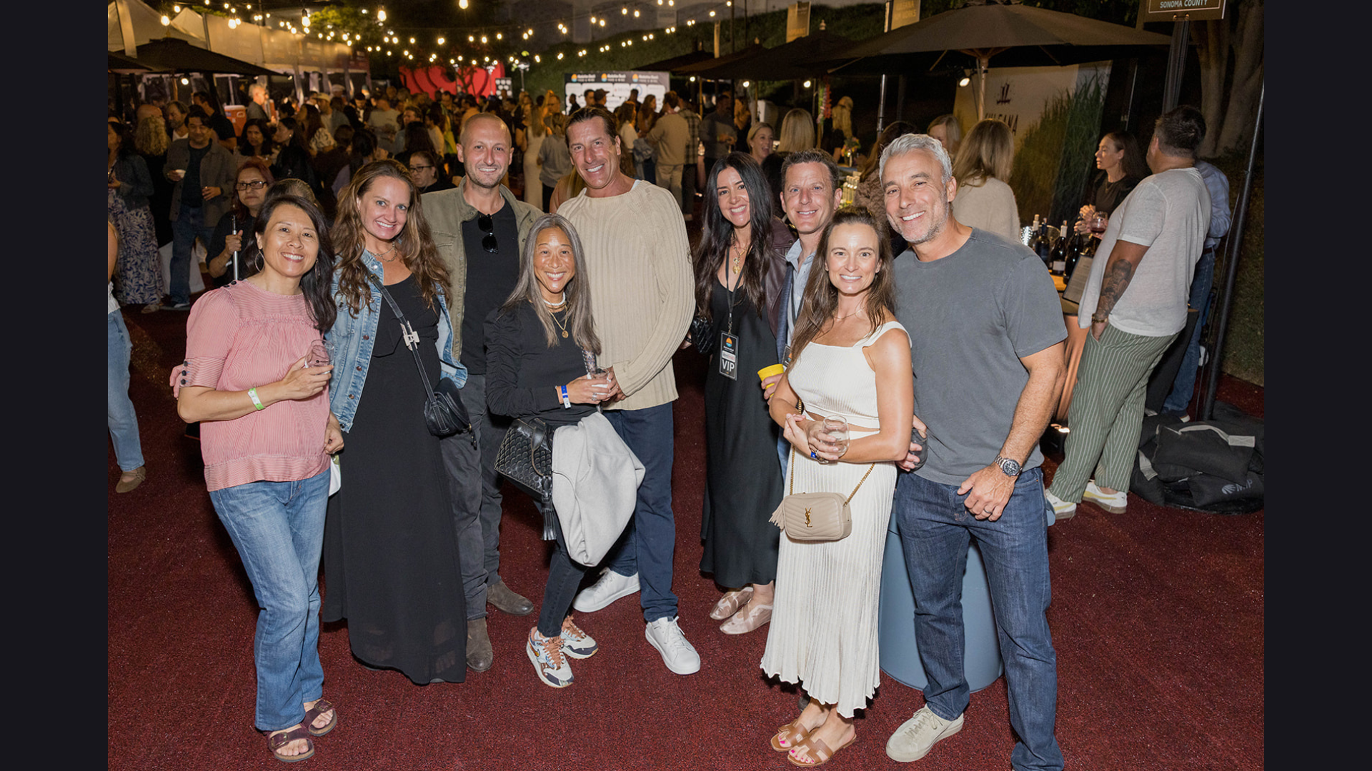 Group of people posing outdoors at a nighttime event with string lights and a red carpet.