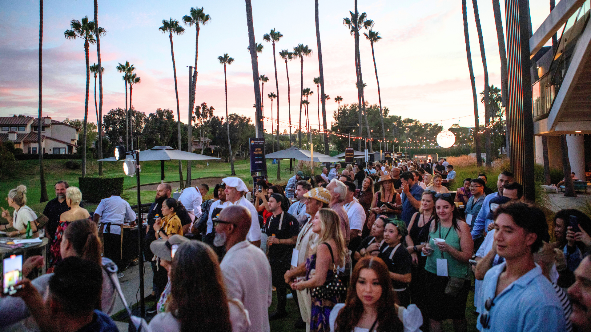 Crowd gathered at an outdoor event at sunset, with palm trees and a building in the background