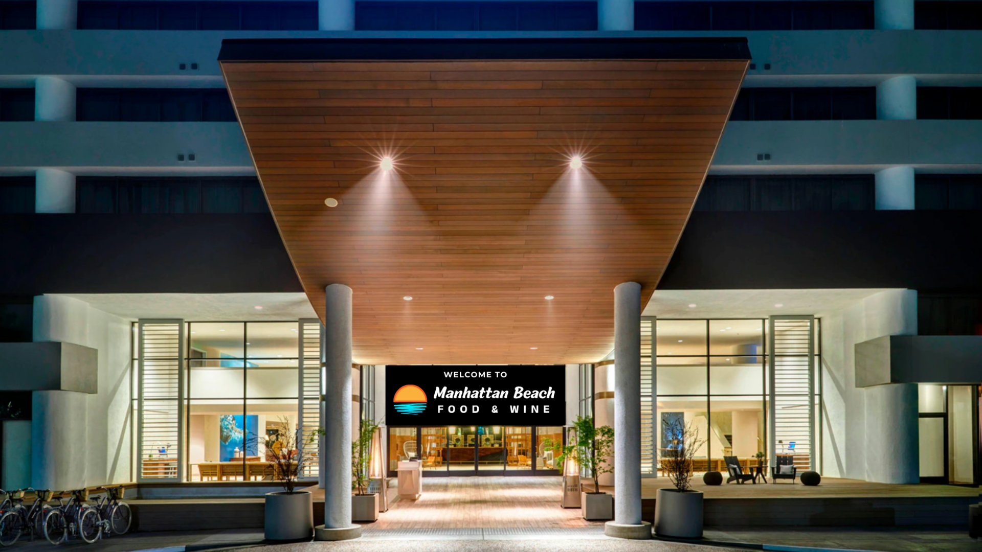 Modern hotel entrance at night with a wood canopy, glass facade, and lit sign reading “Ramada Plaza.”