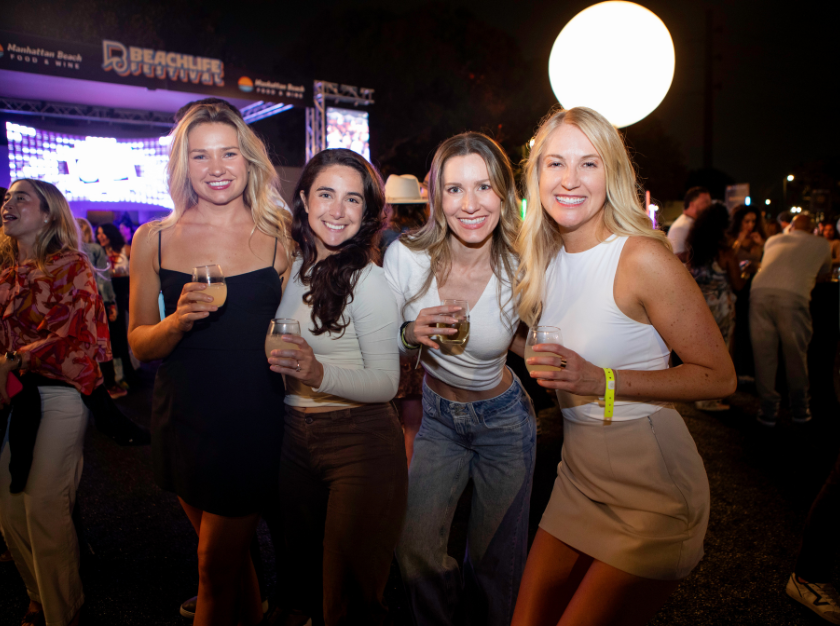 Four women holding drinks at a lively nighttime outdoor event, smiling under bright lights.