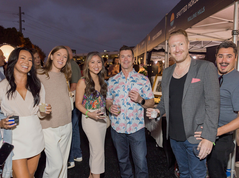 Group of six people socializing outdoors at an evening event, holding drinks under a lit tent.