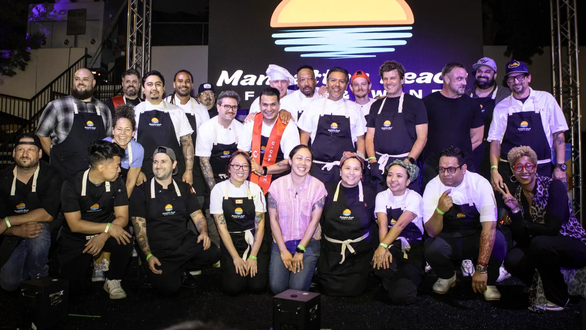 Group of people posing on a stage in front of a lit logo, some wearing black shirts and medals