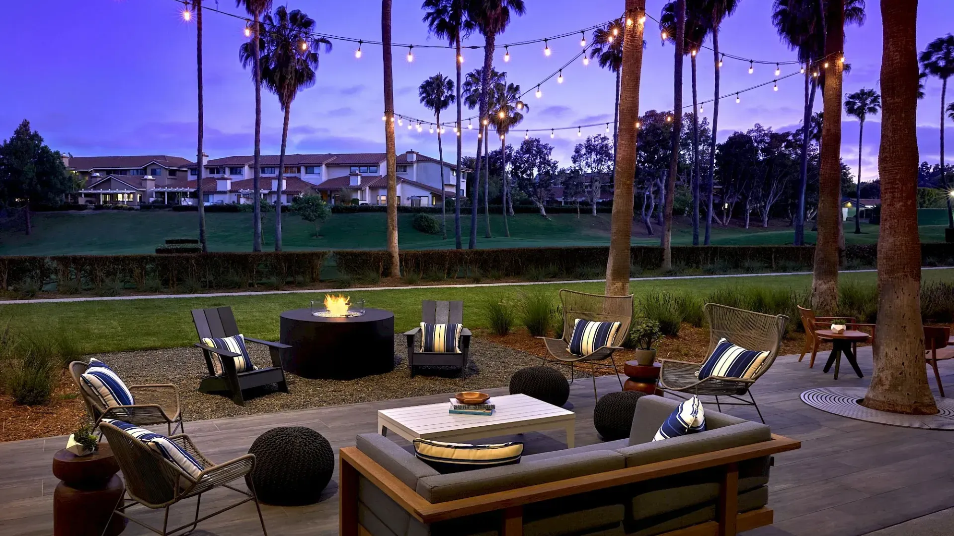 Outdoor patio with lounge chairs around a fire pit at dusk, with string lights and a waterfront view.