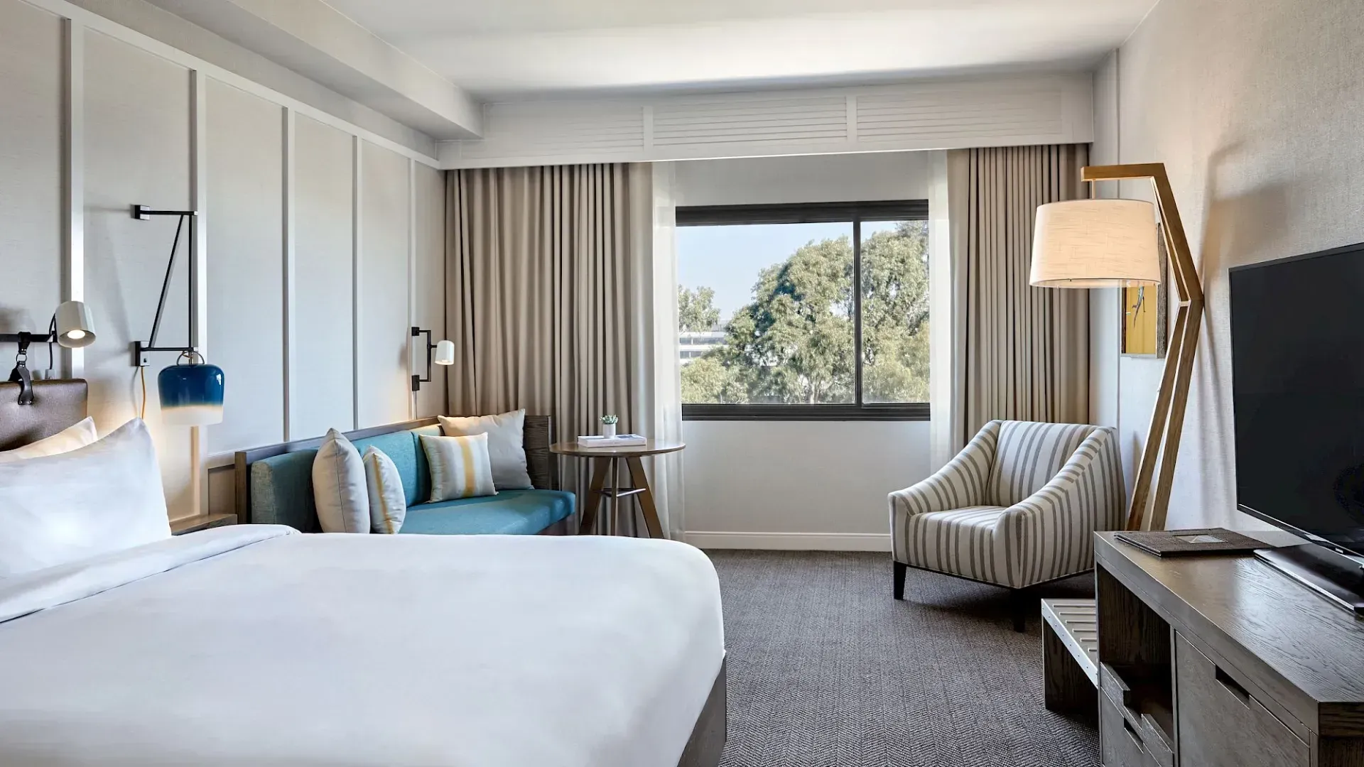 Bright hotel room with a made bed, armchair, desk, window, and lamp.