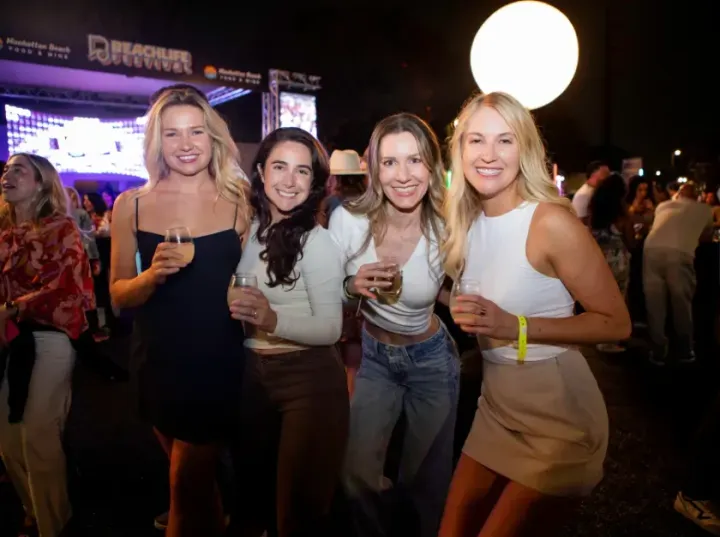 Four women holding drinks at a lively nighttime outdoor party, smiling toward the camera.