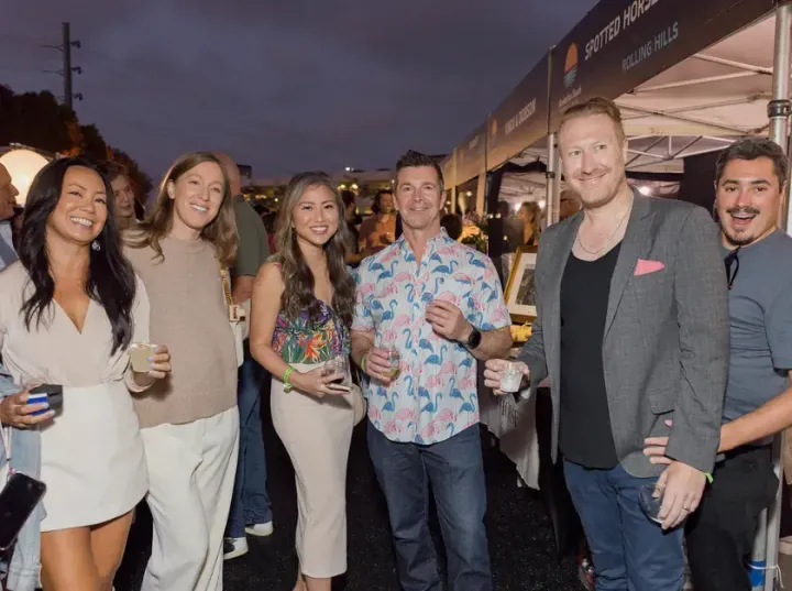 Six people posing with drinks at an outdoor evening event under a tent
