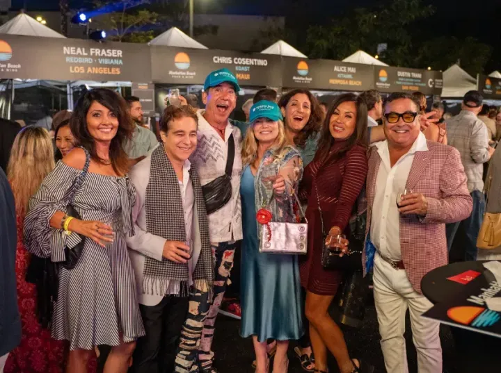 Group of people smiling at an outdoor event at night, standing before vendor tents and holding drinks and awards