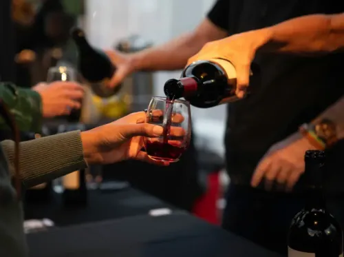 Wine being poured into a glass at a social gathering, with several bottles on a table.