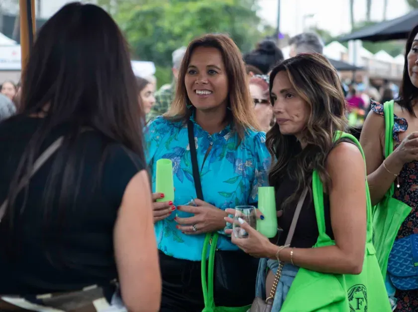 Three women in neon green and blue outfits chatting outdoors at a crowded event.