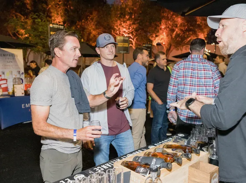 People talking at an outdoor tasting table with bottles and cups at night