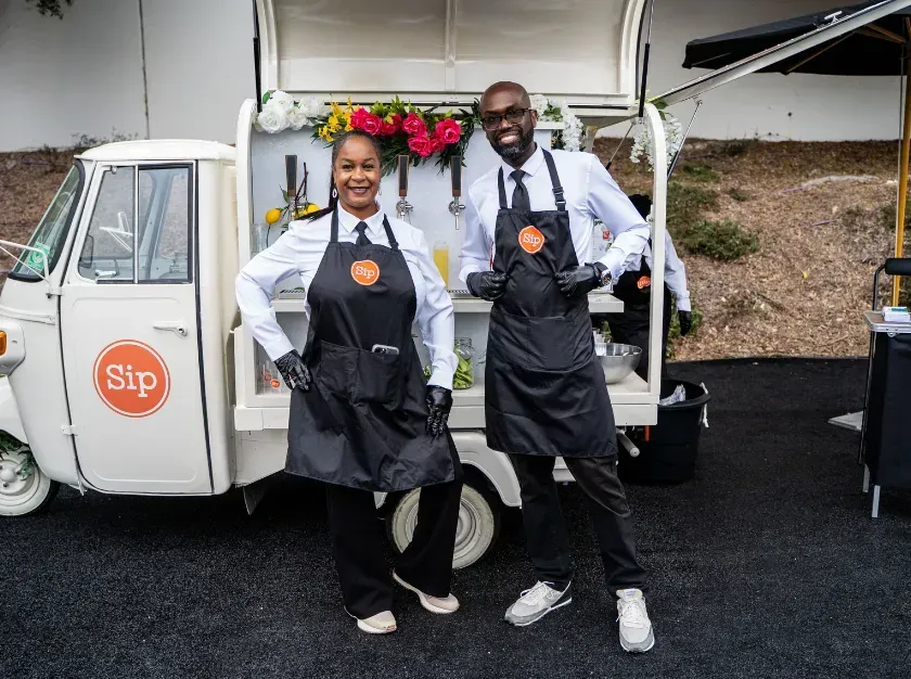 Two smiling vendors in aprons stand beside a Sip delivery van with an open rear hatch.