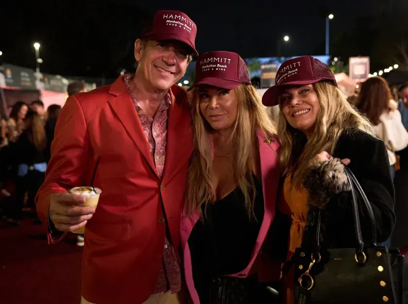 Three people in matching red hats posing at a nighttime event, smiling and holding drinks