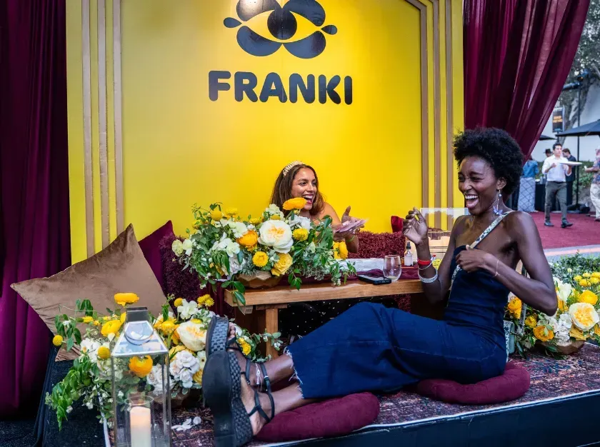Two women posing in front of a yellow FRANKI display with flowers and purple drapes